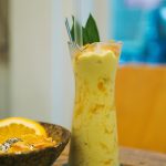 Tall glass of mango smoothie with lemon and straw in a cozy setting.