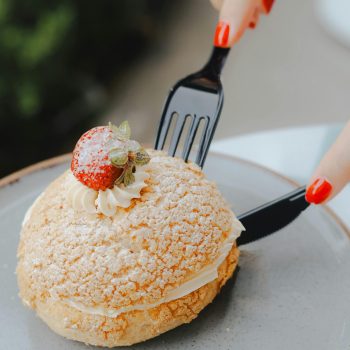 A cream puff topped with a strawberry, being cut with a fork and knife. Captured in Ankara.